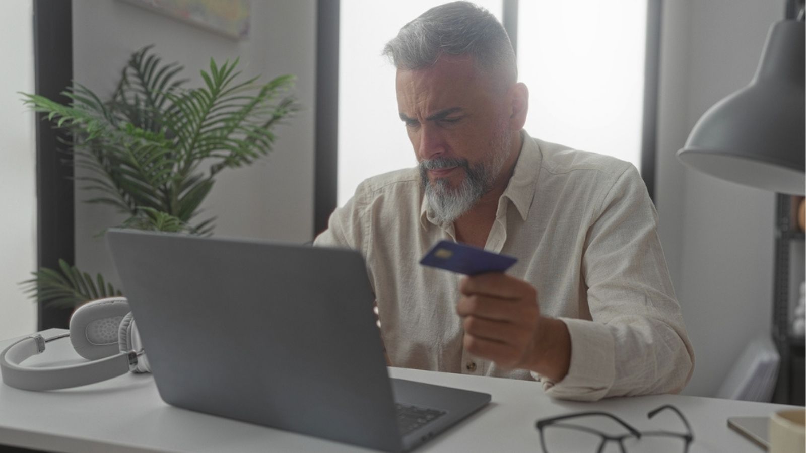 Skupiony mężczyzna w średnim wieku z siwą brodą siedzi przy biurku i patrzy na ekran laptopa, trzymając w ręku kartę płatniczą.